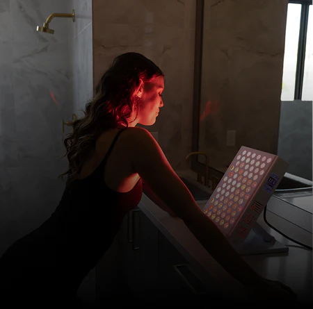 Woman in a tank top looking at red light therapy panel in dark bathroom